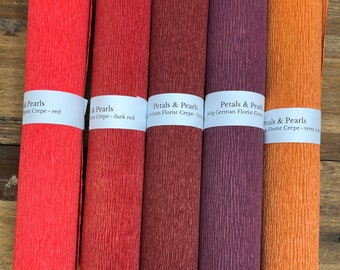 Könnte beinhalten: Fünf Rollen strukturiertes Krepppapier in Rot-, Dunkelrot-, Burgunder-, Lila- und Orangetönen. Jede Rolle ist mit dem Text "Petals & Pearls" und dem Farbnamen beschriftet. Das Papier ist deutsches Floristenkrepp.