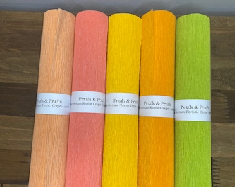May include: Five rolls of colorful crepe paper in shades of peach, coral, yellow, orange, and lime green. Each roll has a white label with the text "Petals & Pearls German Florist Crepe". The paper is textured and appears to be for floral arrangements or crafts.