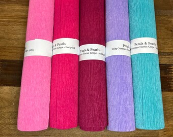 May include: Five rolls of colorful crepe paper in shades of pink, magenta, purple, and teal. Each roll is labeled with the brand "Petals & Pearls" and the color name. The paper has a textured, pleated appearance.