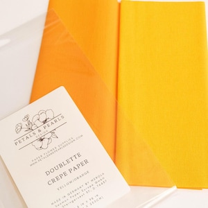 May include: A package of yellow and orange doublette crepe paper by Petals & Pearls Design. The label states the paper is made in Germany by Werola and measures 9.8 inches by 98 inches. The paper is folded and partially visible inside a clear sleeve.