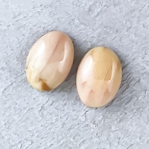 May include: Two oval-shaped gemstones with a polished finish. The stones have a creamy, peach color with subtle variations in tone. They are set against a textured, gray surface, showcasing their natural beauty. These gemstones are ideal for jewelry making.
