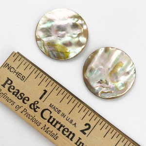 Op de afbeelding: Twee ronde, iriserende parelmoer knopen, ongeveer 2,5 cm in diameter, op een wit oppervlak naast een houten liniaal.