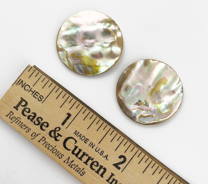 Op de afbeelding: Twee ronde, iriserende parelmoer knopen, ongeveer 2,5 cm in diameter, op een wit oppervlak naast een houten liniaal.