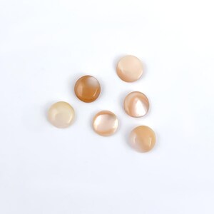 May include: Six round, iridescent buttons in shades of cream, peach, and brown are scattered on a white background. The buttons have a smooth, polished surface and a slight dome shape, suggesting they are made of shell or a similar material.