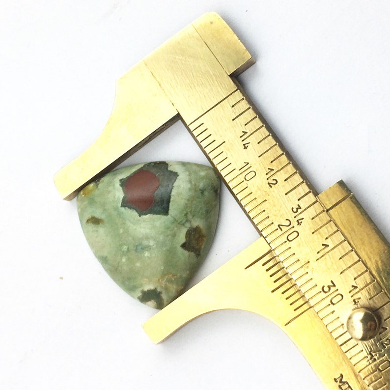 May include: A green, red, and black patterned cabochon gemstone being measured with a brass caliper. The gemstone is approximately 1.25 inches wide.