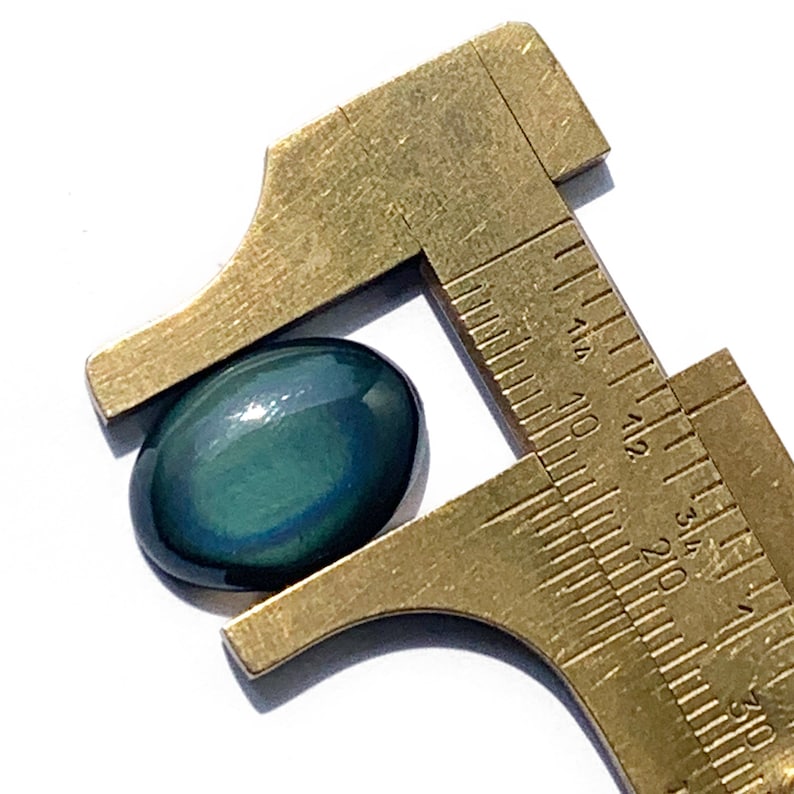 May include: A dark green oval gemstone with iridescent blue and purple hues is measured with a caliper. The gemstone is approximately 1.5 inches long.