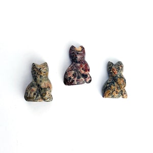 May include: Three small, intricately carved stone cat figurines. The cats are in various poses and have a mottled brown, gray, and red color scheme.