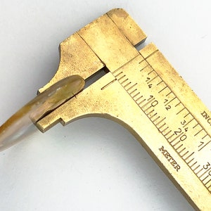 Op de afbeelding: Een messing schuifmaat met een wit en bruin schelpvormig object dat wordt gemeten. De schuifmaat heeft markeringen in inches en millimeters.