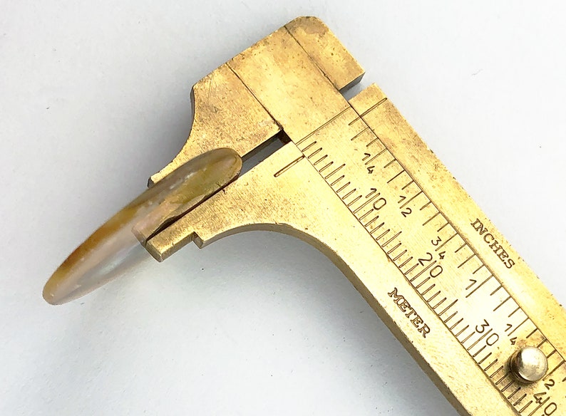 Op de afbeelding: Een messing schuifmaat met een wit en bruin schelpvormig object dat wordt gemeten. De schuifmaat heeft markeringen in inches en millimeters.