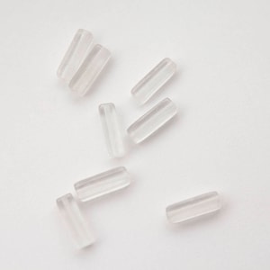 Op de afbeelding: Een groep van heldere glazen kralen, elk gevormd als een klein, rechthoekig prisma. De kralen zijn verspreid over een wit oppervlak.