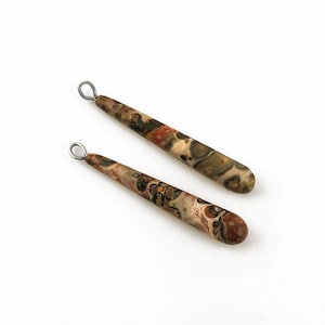 Peut inclure: Deux pendentifs en pierre en forme de goutte d'eau avec un motif tacheté dans les tons de marron, beige et rouge. Chaque pendentif a une petite boucle argentée en haut. Les pendentifs sont présentés sur un fond blanc.