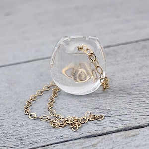 May include: A clear, spherical pendant with a gold-colored chain. The pendant has a small, gold-colored object inside. The chain is delicate and hangs down from the pendant. The pendant is sitting on a light gray surface.