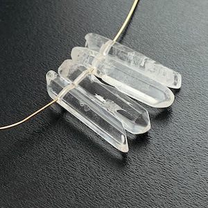 May include: Clear quartz crystal pendant with a gold-colored wire. The crystals are elongated and faceted, with a natural, translucent appearance. The pendant is displayed against a dark background.