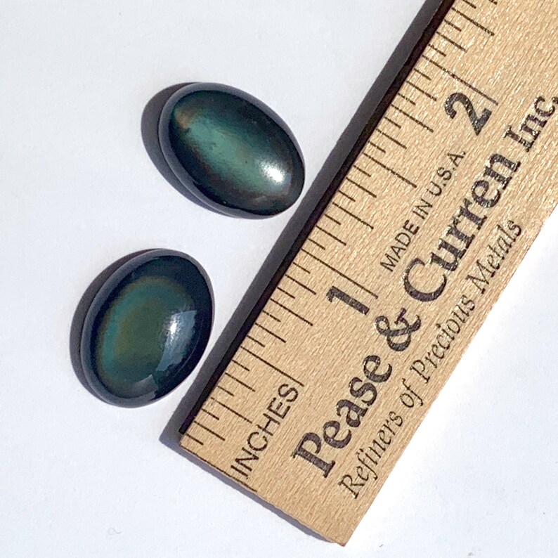 May include: Two oval-shaped, iridescent green and black stones, each approximately 1 inch long, are displayed on a white surface next to a wooden ruler. The ruler is marked in inches and has the text "Pease & Curren Inc. Refiners of Precious Metals" printed on it.