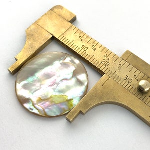 Op de afbeelding: Een messing schuifmaat meet een ronde, iriserende parelmoerknop. De knop heeft een diameter van ongeveer 2,5 cm.
