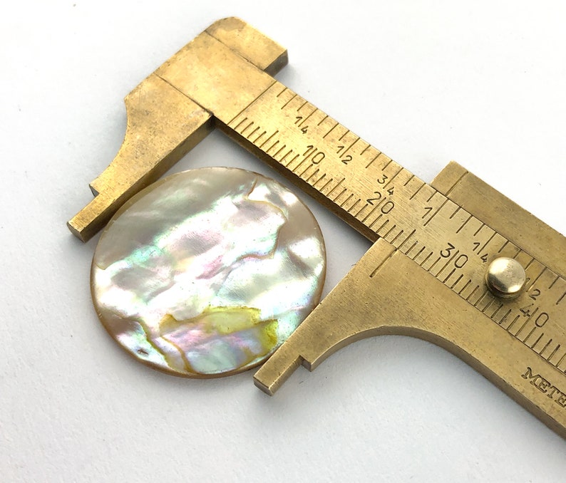 Op de afbeelding: Een messing schuifmaat meet een ronde, iriserende parelmoerknop. De knop heeft een diameter van ongeveer 2,5 cm.