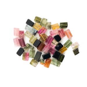 Può includere: Una collezione di pietre preziose sfaccettate rettangolari e colorate. Le pietre preziose sono di varie tonalità di rosa, verde, nero e marrone. Le pietre preziose sono disposte in un mucchio su uno sfondo bianco.