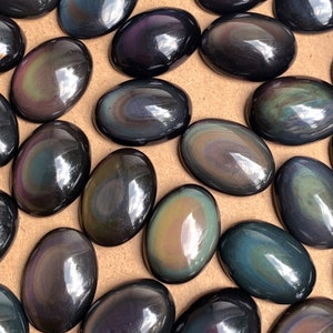 May include: A collection of oval-shaped obsidian stones with iridescent rainbow colors. The stones are smooth and polished, and they are arranged in a random pattern on a brown surface.