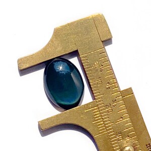 May include: A dark green oval gemstone with a metallic sheen is being measured with a brass caliper. The caliper is marked in millimeters and the gemstone measures approximately 17 mm in length.
