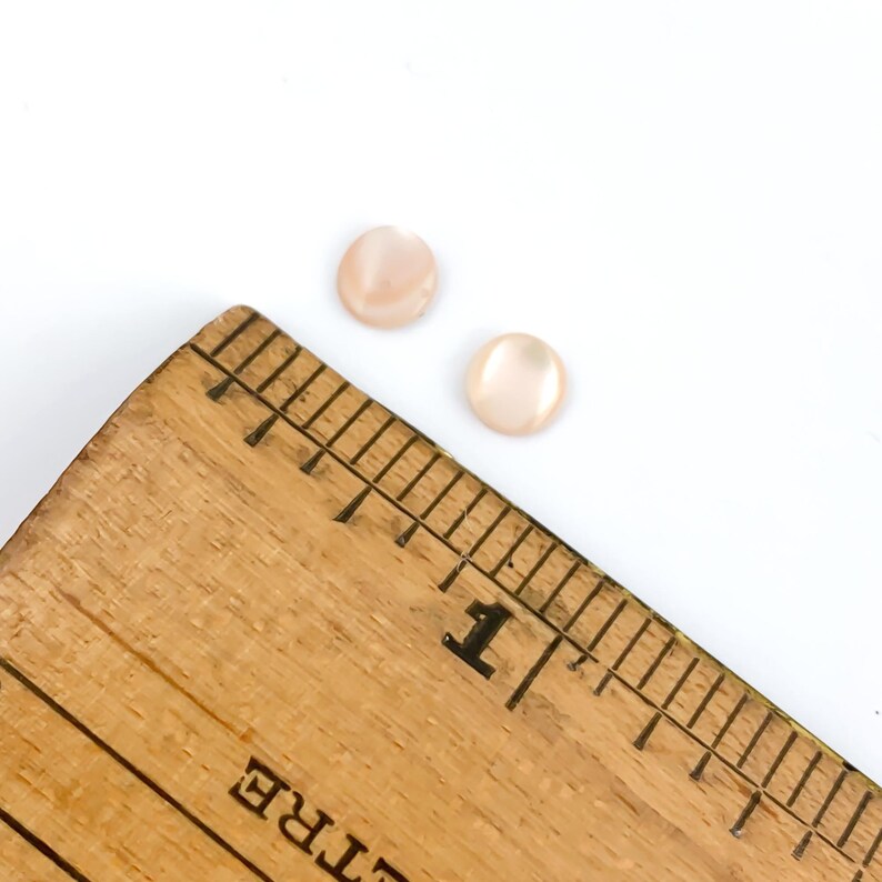 May include: Two small, round, pale pink buttons are displayed on a white background. A wooden ruler with black markings and the number 1 is in the lower left corner. The buttons appear to be approximately 0.5 inches in diameter.