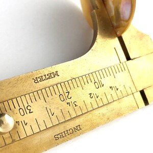 Op de afbeelding: Een meetinstrument van messing met een schuifschaal die in inches en centimeters is gemarkeerd. De schaal is ge&euml;tiketteerd met "INCHES" en "METER".