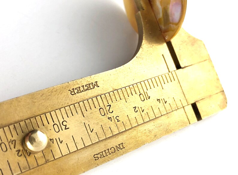 Op de afbeelding: Een meetinstrument van messing met een schuifschaal die in inches en centimeters is gemarkeerd. De schaal is ge&euml;tiketteerd met "INCHES" en "METER".