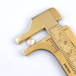 May include: A brass caliper measuring a small, pale pink, round object. The caliper is marked with inch measurements. The object appears to be a cabochon or a similar small, rounded component, possibly for jewelry or crafts.