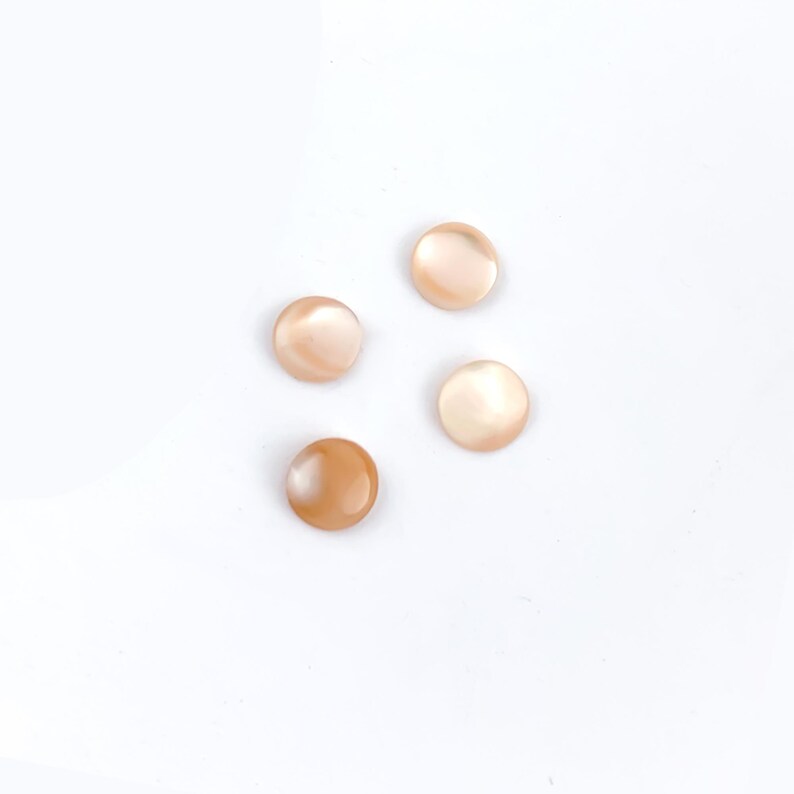 May include: Four round, iridescent buttons in shades of peach and tan are scattered on a white background. The buttons have a smooth, polished surface and a subtle, pearlescent sheen, suggesting they are made of shell or a similar material.