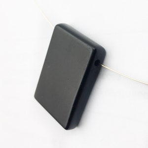 Puede incluir: Un colgante rectangular negro con un agujero para un cordón o cadena. El colgante está hecho de piedra negra pulida.