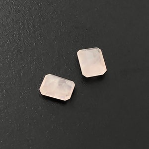 May include: Two pale pink rectangular gemstones with a smooth, polished finish. The gemstones are set against a black background.