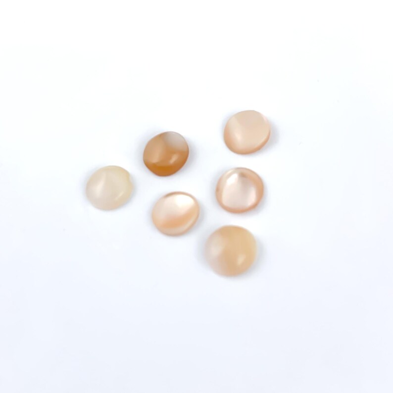 May include: Six oval-shaped, polished gemstones in varying shades of peach and cream are scattered on a white surface. The stones have a smooth, glossy appearance, with subtle color variations. The gemstones are likely cabochons, suitable for jewelry making or crafts.