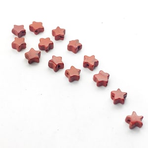 May include: A collection of 14 red star-shaped beads made of red jasper. The beads are arranged in a curved line on a white background.