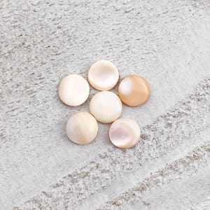 May include: Six round, polished, shell buttons in shades of cream and peach are arranged on a textured, light gray surface. The buttons have a subtle iridescent sheen, reflecting light. These buttons are ideal for sewing and craft projects.