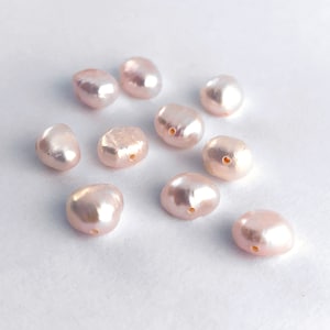 Peut inclure: Une collection de dix perles d'eau douce rose pâle avec un trou percé au centre de chaque perle. Les perles sont de forme ovale et ont une finition lisse et brillante.