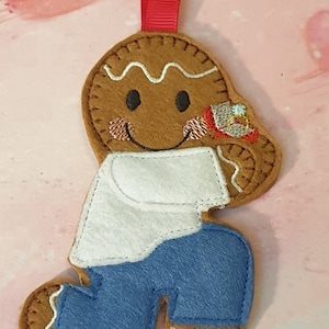 May include: A brown felt gingerbread person ornament with a red ribbon loop. The figure wears a white shirt and blue trousers, has stitched facial features with pink cheeks, and holds a small red box containing a ring with a blue stone.