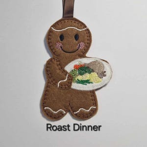 May include: A brown felt gingerbread man decoration, smiling and holding a plate with a roast dinner. It has a brown ribbon for hanging and the text "Roast Dinner" below.