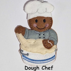 May include: A felt ornament depicting a chef with brown skin, wearing a white chef's hat and grey uniform. The chef holds a bowl of dough. The bowl is white with blue stripes. The text "Dough Chef" is below the image.