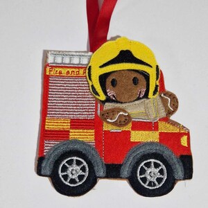 May include: A felt ornament shaped like a red fire engine with yellow checkered accents, black wheels, and a brown gingerbread character wearing a yellow firefighter helmet. The engine features a red ribbon hanger and the text Fire and Rescue.