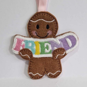 May include: A gingerbread person ornament with a smiling face, holding a sign that says "FRIEND" in rainbow colours.