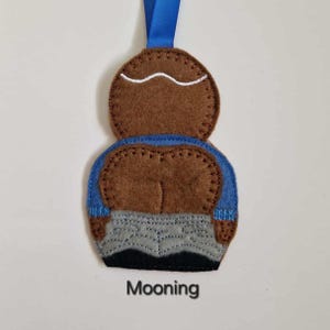 May include: A handmade felt ornament depicting a person mooning. The ornament is brown, blue, grey, and black, with a blue ribbon for hanging. The word "Mooning" is printed below the ornament.