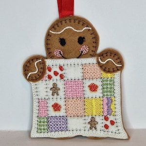 May include: A gingerbread man ornament holding a patchwork quilt with red, pink, yellow, green, and purple squares. The quilt has gingerbread men and red dots embroidered on it.