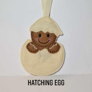 May include: A cream-coloured felt ornament depicting a hatching egg. A gingerbread man with a smiling face and brown details is emerging. The ornament has a cream-coloured ribbon for hanging, and the text "HATCHING EGG" is printed below.
