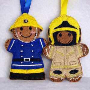 May include: Two felt ornaments shaped like gingerbread people dressed as firefighters. One wears a yellow helmet and blue uniform with a black belt. The other wears a yellow and black helmet with a tan turnout coat featuring yellow reflective stripes.