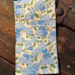 May include: A rectangular paper napkin featuring a repeating pattern of blue hydrangea flower clusters and green leaves on a white background. The napkin is folded and displays a detailed botanical print with various shades of blue and green.