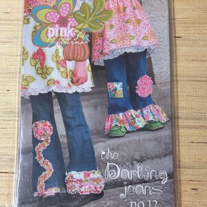 May include: A sewing pattern packet featuring a design for flared jeans with floral and ruffle embellishments. The packet has the text "pink fig patterns" and "the Darling jeans no.12". The jeans are shown with a floral top.