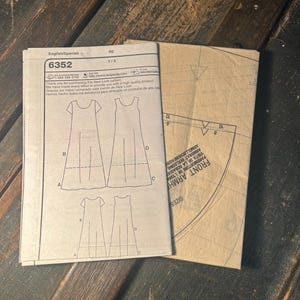 May include: A New Look sewing pattern envelope labeled 6352, featuring line drawings of a sleeveless dress with variations labeled A, B, C, and D. Beside it lies a folded brown paper pattern piece with printed text reading FRONT ARMHOLE.