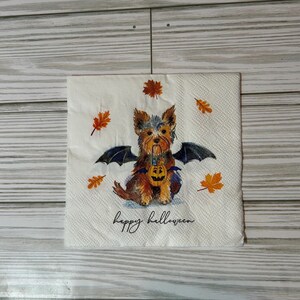 May include: Square white paper napkin with a Halloween-themed design. The illustration features a Yorkshire Terrier with bat wings, holding a jack-o'-lantern. The words "happy halloween" are written in a script font, with autumn leaves scattered around.