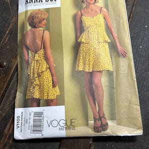 May include: Vogue Patterns Anna Sui dressmaking pattern. The pattern features a yellow dress with a floral print. The front of the pattern shows a woman wearing the dress. The back of the pattern shows a woman wearing the dress. The pattern size is DD (12-14-16-18).