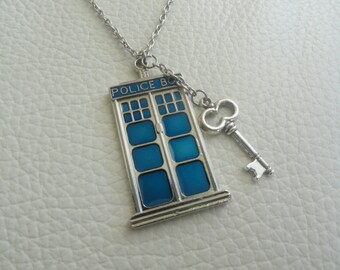 May include: A silver-toned metal pendant necklace featuring a rectangular blue police box charm with the text POLICE BO visible at the top, accompanied by a small silver-toned skeleton key charm attached to the same link on the chain.