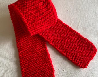 May include: A bright red, hand-knitted scarf. The scarf is made of a thick yarn, with a visible knit pattern. The scarf is folded over on itself, creating a loop. The scarf is lying on a white surface.